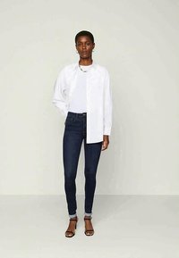 Bela srajca s kratkimi rokavi, prepletena čez oprijet bel T-shirt, v kombinaciji s temno modrimi oprijetimi jeans hlačami in črnimi sandali na peto. Prikazani so minimalni dodatki.