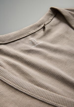Világosbarna bordázott póló kerek nyakkivágással, finom márkajelzéssel a gallér közelében. Az anyag puhanak és texturáltnak tűnik.