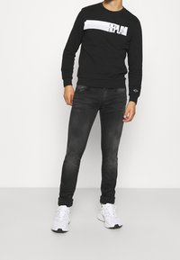Sudadera negra con un logo de rayas blancas en la parte frontal, combinada con jeans oscuros ajustados y zapatillas deportivas blancas.