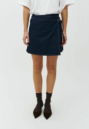 Frau trägt einen dunklen Jeans-Wickelrock mit orangefarbenen Nähten, ein weißes kurzärmeliges Oberteil, dunkle Socken und spitze schwarze Schuhe.