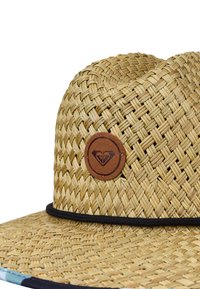 Chapeau en paille tressée à large bord, avec un logo rond en patch de cuir marron et une fine bande noire autour de la base de la calotte.