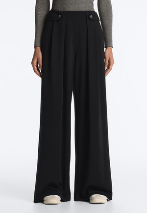 Pantaloni negri cu picioare largi, pliseuri frontale și buzunare cu clapetă și nasturi, purtați cu adidași bej și un top gri cu mâneci lungi, cu model striat.