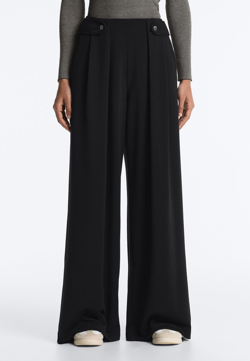 Pantalon noir à jambes larges avec des plis sur le devant et des poches à rabat boutonnées, porté avec des baskets beiges et un haut gris côtelé à manches longues.