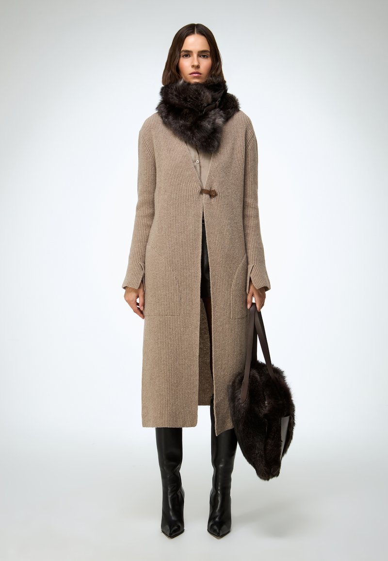 Beežist kudumiga pikk kardigan, millel on nööpkinnitused, kombineeritud tumeda karvaga salliga ja hoides pruuni karvaga kotti. Mustad põlvikukõrgused saapad.