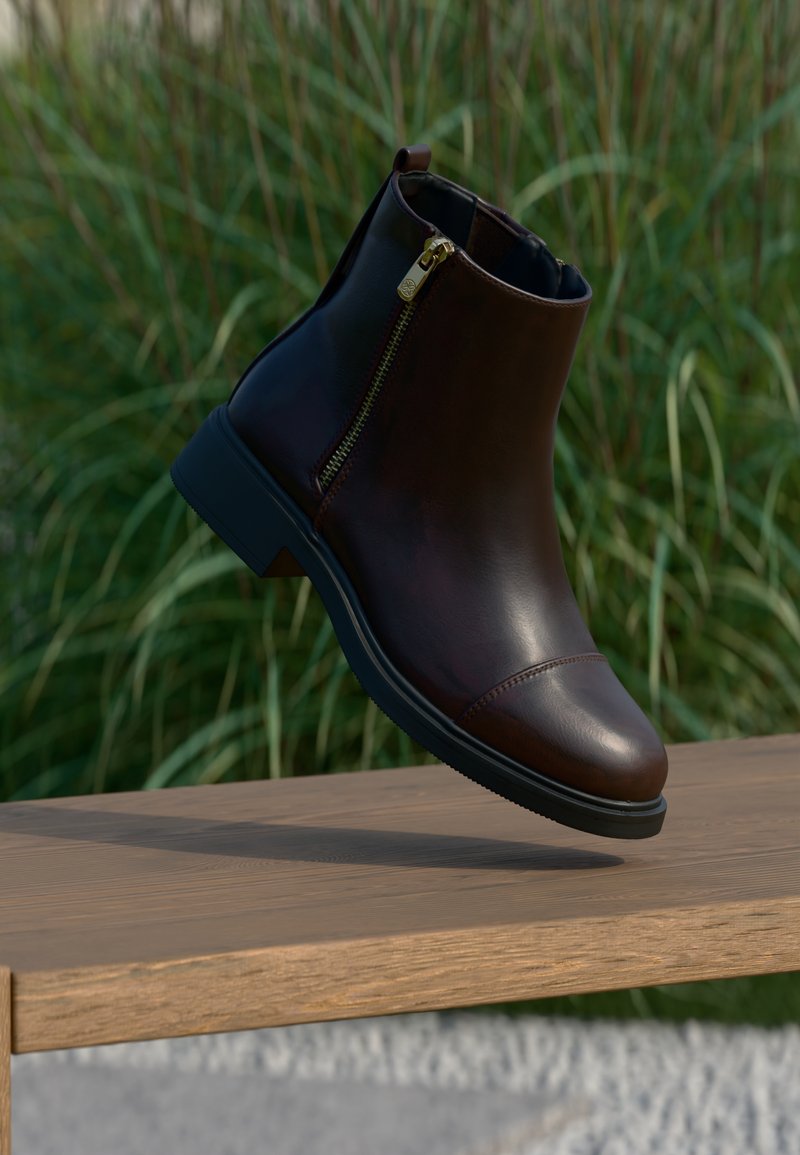 Bota de cano curto em couro castanho com biqueira curva, detalhe de fecho dourado e sola de borracha preta, repousando sobre uma superfície de madeira.