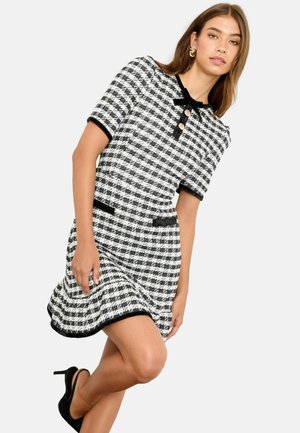 Musta-valkoinen tweed-mekko lyhyillä hihoilla, pyöreällä kauluksella, mustalla reunuksella, kolmella napilla ja kahdella etutaskulla; liehuva helma.
