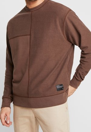 Uomo che indossa una felpa a maniche lunghe color marrone con una piccola etichetta nera sul lato inferiore, abbinata a pantaloni beige.