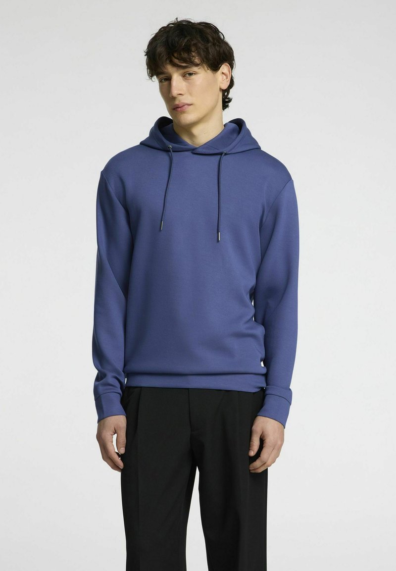 Felpa viola con cappuccio e cordini, maniche lunghe e vestibilità comoda, abbinata a pantaloni neri. Design minimalista, texture liscia.