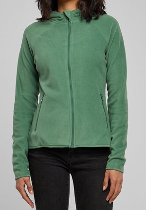 Femme portant une veste polaire verte zippée avec poches latérales zippées et un pantalon noir sur fond gris.