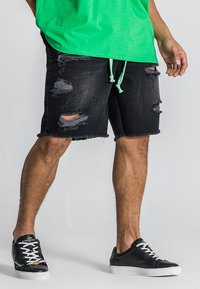 Pantalones cortos de mezclilla negra con detalles desgastados y dobladillos deshilachados, combinados con zapatillas negras con suelas blancas y detalles de cordones.