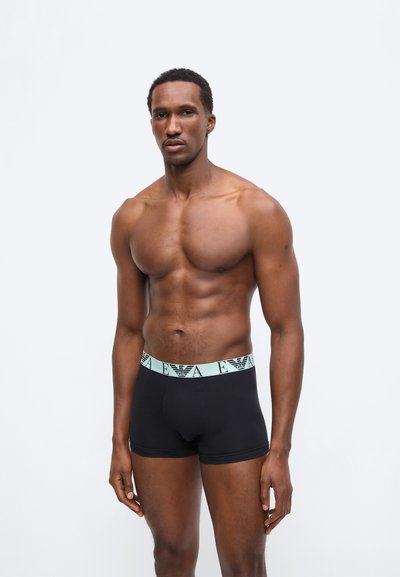 Μυώδης άνδρας που στέκεται μπροστά σε λευκό φόντο, φορώντας μαύρα αντρικά εσώρουχα με ανοιχτό μπλε ζωνάκι στη μέση που φέρει λογότυπο.