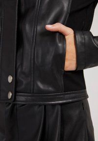Veste en cuir noir avec une texture lisse, des boutons-pression et une poche cachée. Le design associe cuir et tissu pour un contraste.