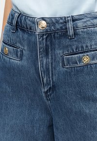 Τζιν παντελόνια σε σκούρο μπλε χρώμα με ελαφρώς ξεθωριασμένη υφή. Διαθέτουν χρυσούς μεταλλικούς εξαρτήματα και εμπρόσθιες τσέπες με καπάκια.