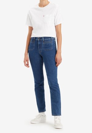 Person wearing a white short-sleeve pocket t-shirt, high-waisted blue jeans, white socks, and white sneakers standing against a white background.