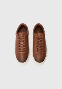 Zapatillas de cuero marrón con puntera redonda, cordones planos y suela de goma blanca. Presenta un logotipo sutil en la lengüeta y el costado.