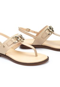 Sandalias de cuero beige con diseño tipo thong, adornadas con cadenas en tono dorado y correas ajustables en el tobillo. Textura suave y tacón bajo.