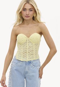 Femme blonde portant un corset bustier en dentelle jaune et un jean bleu clair décoré de perles blanches le long des côtés et des poches.