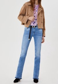 Mörk suède kort jacka med dubbelknäppt design, rosa blommig topp under, ljusblå jeans, svart bälte och svarta klackade ankelstövlar.