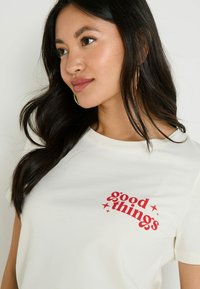 T-shirt en coton blanc avec un graphique rouge portant le texte "bonnes choses" orné d'étoiles, à manches courtes et à col rond.