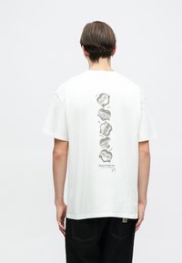 Biała bawełniana koszulka z nadrukiem na plecach, przedstawiająca szereg etykiet oraz nazwę marki "Carhartt". Krótkie rękawy i standardowy krój.