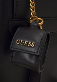 Detail černé kožené kabelky se zlatým řetízkem a malou černou taštičkou s logem "GUESS" ve zlatém písmu.
