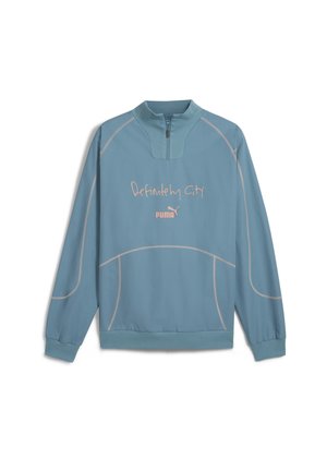 Lyseblå pullover med halv lynlås krave. Har pink broderet tekst "Definitely City" og et lille logo. Glat, letvægtsstof.