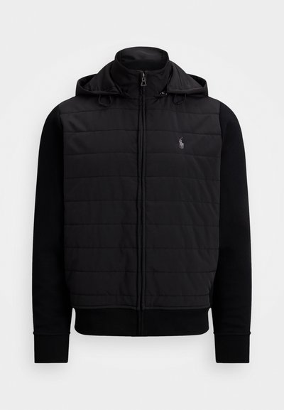 Svart jakke med polstret front, halvglidelås og hette. Har kontrasterende glatte svarte ermer og en liten logo på brystet.