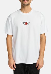 Camiseta de algodón blanca con cuello redondo, que presenta un logo de RVCA en negro y rojo impreso en el pecho. Manga corta, corte relajado.