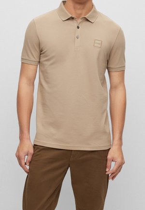 Beige Poloshirt mit klassischem Kragen, zwei Knöpfen und einem kleinen quadratischen Logo auf der Brust. Kurze Ärmel und eine glatte, weiche Textur.