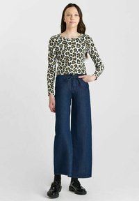 Langarm geribbelde top met een groen en zwart luipaardpatroon, gecombineerd met donkerblauwe wijde broek en zwarte schoenen.