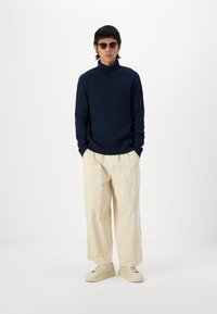 Suéter de cuello alto de canalé en color navy, pantalones de pierna ancha en crema y zapatillas de plataforma beige, con gafas de sol redondas que completan el conjunto.