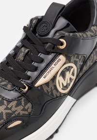 Baskets Michael Kors noires et dorées avec tissu à motifs, lacets noirs, accents brillants en cuir verni et logo sur le côté et la languette.