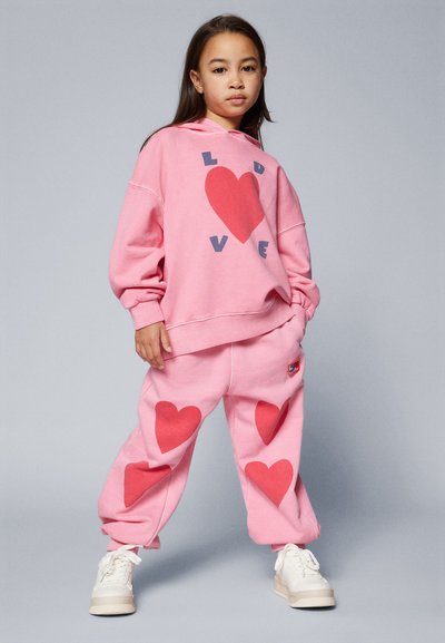 Rózsaszín kapucnis pulóver "LOVE" grafikával és piros szível, rózsaszín melegítőnadrággal, nagy piros szívmintákkal párosítva. A megjelenést fehér sportcipők teszik teljessé.