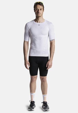 Homme debout face à l'avant, portant un maillot de compression blanc, un short de cyclisme noir, des chaussettes blanches de marque, des chaussures de course noires et des lunettes de soleil de sport jaunes.