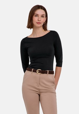 Vrouw draagt een aansluitend zwart driekwart mouw topje, beige broek met hoge taille en een bruine riem met een gouden gesp, staand tegen een witte achtergrond.