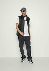 Jonge man met een groene pet, zwarte zonnebril, donkergroene gewatteerde vest, witte t-shirt met logo, losse zwarte spijkerbroek en witte sneakers.