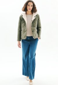 Nainen kihara hius, päällä vihreä peitettävä takki, jossa on fleecevuori, vaaleanbeige paita, siniset leveälahkeiset farkut ja vaaleanbeige teräväkärkiset kengät.