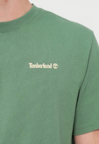Πράσινο βαμβακερό μπλουζάκι με στρογγυλή λαιμόκοψη και κοντά μανίκια. Το όνομα της μάρκας "Timberland" και το λογότυπο είναι τυπωμένα σε κρεμ χρώμα στο στήθος.