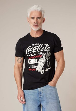 Man met wit haar en baard, gekleed in een zwart Coca-Cola T-shirt met bezorgafbeelding en lichtblauwe spijkerbroek, staand tegen een effen achtergrond.
