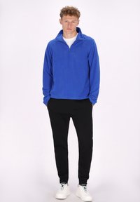 Pullover in felpa blu con colletto alto e zip a quarto, abbinato a pantaloni della tuta neri a sigaretta e scarpe da ginnastica bianche. Texture liscia, design casual.