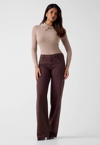 Beige geribbelde top met lange mouwen en knoopdetails, gecombineerd met bruine uitlopende spijkerbroek. De outfit heeft een aansluitende pasvorm en een gladde textuur.