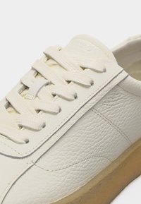 Λευκό δερμάτινο αθλητικό παπούτσι με υφή στην επιφάνεια, λευκά κορδόνια και ανοιχτό καφέ ελαστική σόλα, εμφανιζόμενο σε κοντινή λήψη από το πλάι και από πάνω.