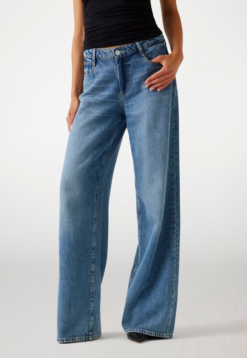 Personne portant un jean bleu taille haute à jambes larges et un top noir sans manches, debout avec une main dans la poche et chaussures noires pointues visibles.