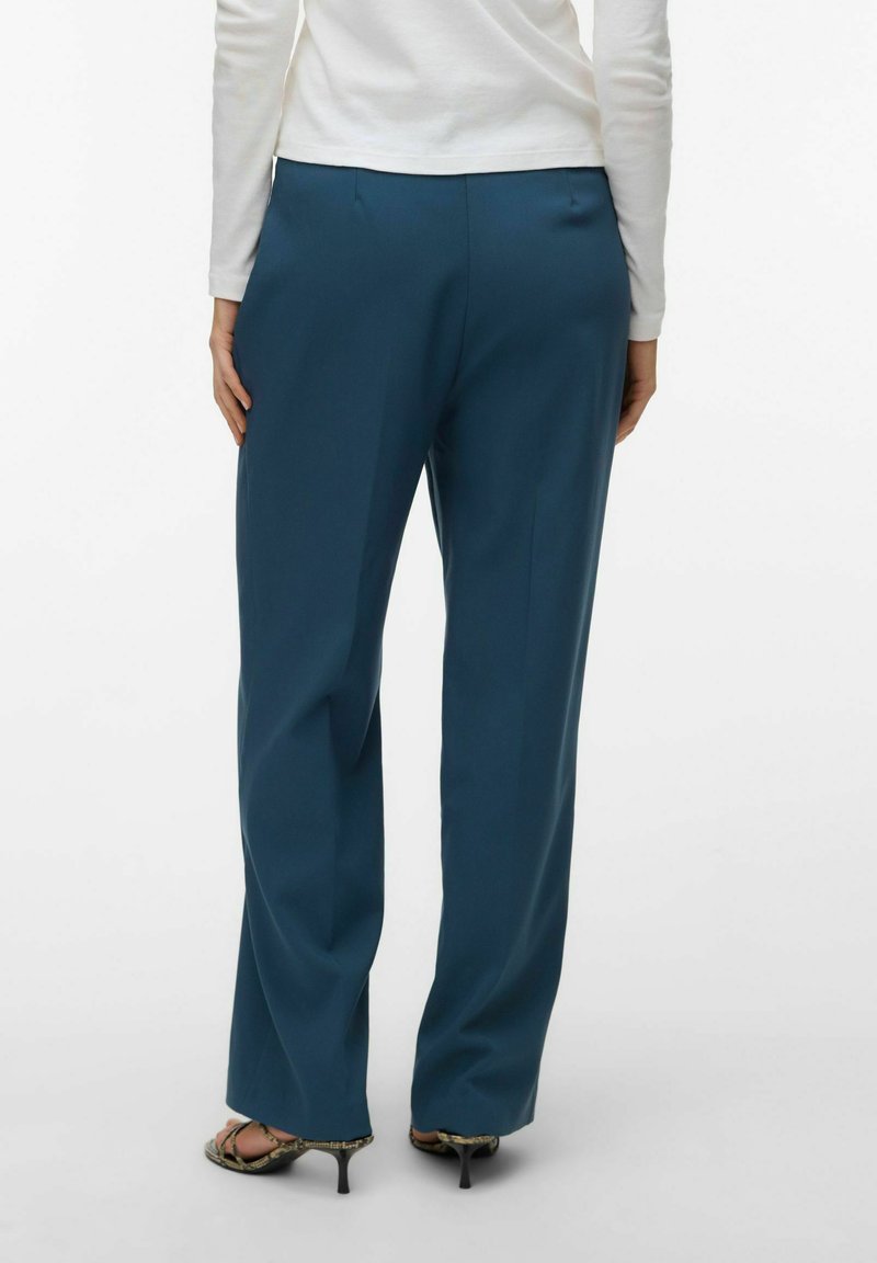 Femme portant un pantalon large bleu sarcelle foncé, un haut blanc à manches longues et des sandales à talons ouverts, debout de dos sur un fond uni.