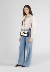 Borsa a tracolla; pelle bianca e nera; forma rettangolare; accessori in argento; modello indossa una camicia marrone chiaro con colletto arricchito e jeans larghi blu.