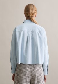 Helehall, lühendatud pikkade varrukatega nööbitav särk terava kaelusega, kombineeritud ruuduliste pükstega. Materjal tundub sile, ilma tekstuurita.