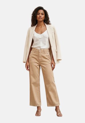 Donna con pantaloni beige a gamba larga con dettagli a bottoni, canotta bianca in raso, blazer crema sulle spalle e sandali con cinturini.