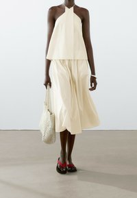 Haut halter beige clair et jupe fluide assortie en tissu texturé. Femme portant un sac tissé crème et des tongs avec des accents rouges.