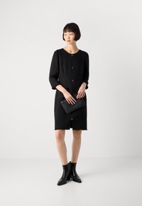 Abito nero, texture, con maniche a tre quarti, scollatura tonda e cinque bottoni sul davanti; abbinato a una pochette nera e stivaletti.