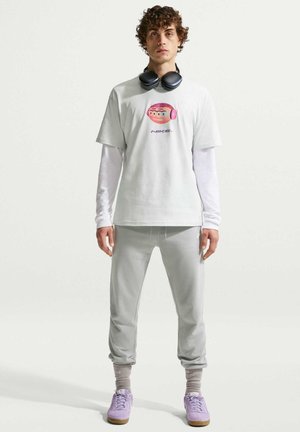 Młody mężczyzna w białej koszuli z długim rękawem pod białym graficznym t-shirtem, jasnoszarych spodniach dresowych, fioletowych trampkach i czarnych słuchawkach na szyi, stojący.